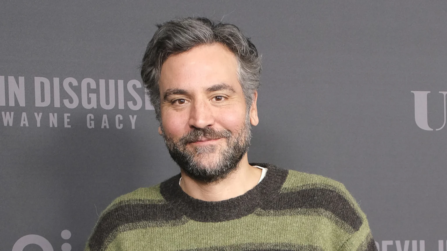 Josh Radnor, with graying hair and a beard, smiles slightly while wearing a green and dark striped sweater against a gray background with faint text.