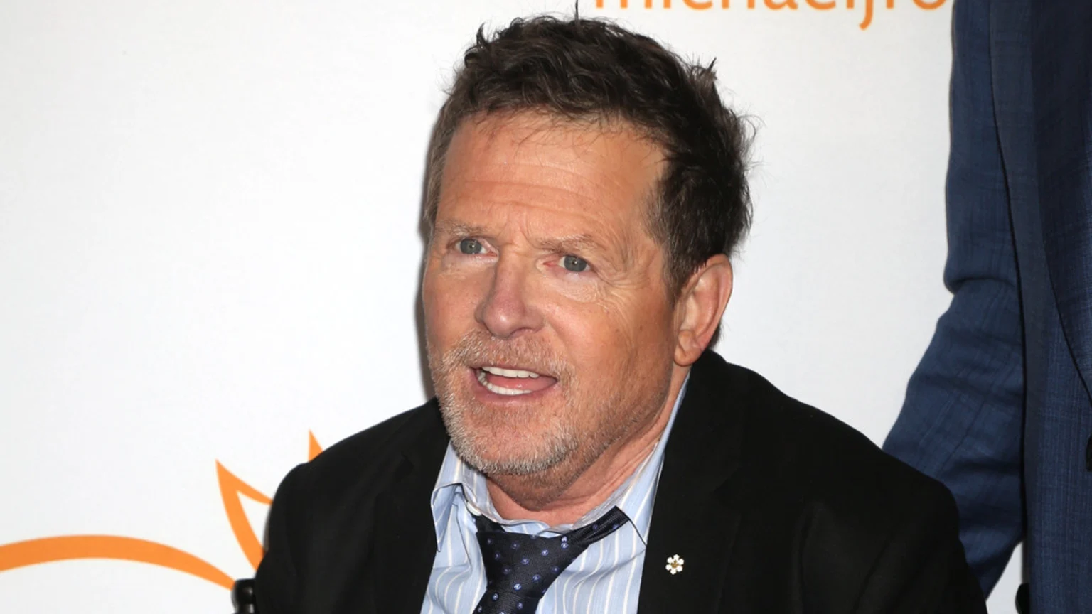 A close-up of actor Michael J. Fox smiling, wearing a black suit jacket over a striped shirt and polka-dot tie, with a white flower pin on his lapel.
