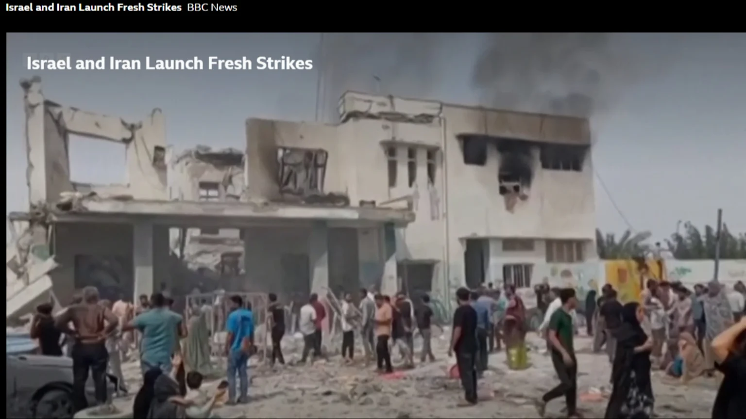 A crowd of people stands amidst rubble in front of heavily damaged buildings, one with smoke rising, with the on-screen text "Israel and Iran Launch Fresh Strikes".