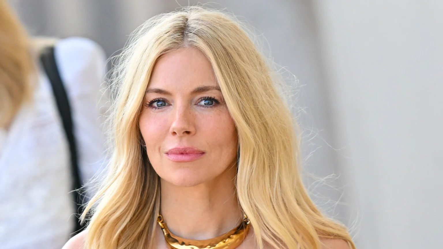 A close-up portrait of a woman with long blonde hair and blue eyes, wearing light makeup and a gold statement necklace, looking directly at the viewer.