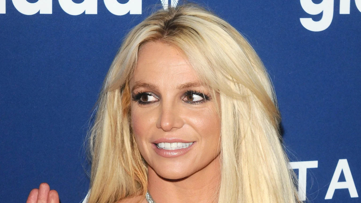 Smiling Britney Spears with long blonde hair and dark eye makeup looks to her right against a blue background with white letters.