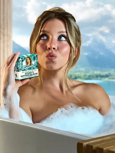 Sydney Sweeney in a bathtub holding up a bar of soap infused with her bathwater.