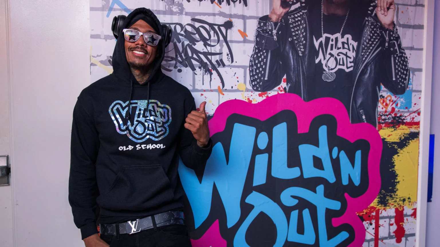 Nick Cannon smiles, wearing a black hoodie with a "Wild 'N Out Old School" logo, star-shaped sunglasses, and headphones, giving a thumbs-up in front of a colorful graffiti-style "Wild 'N Out" backdrop.