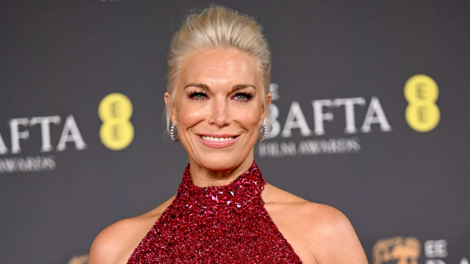 Hannah Waddingham smiles directly at the camera, wearing a sparkling red halter-neck gown and large earrings, with the blurred BAFTA Film Awards logo behind her.