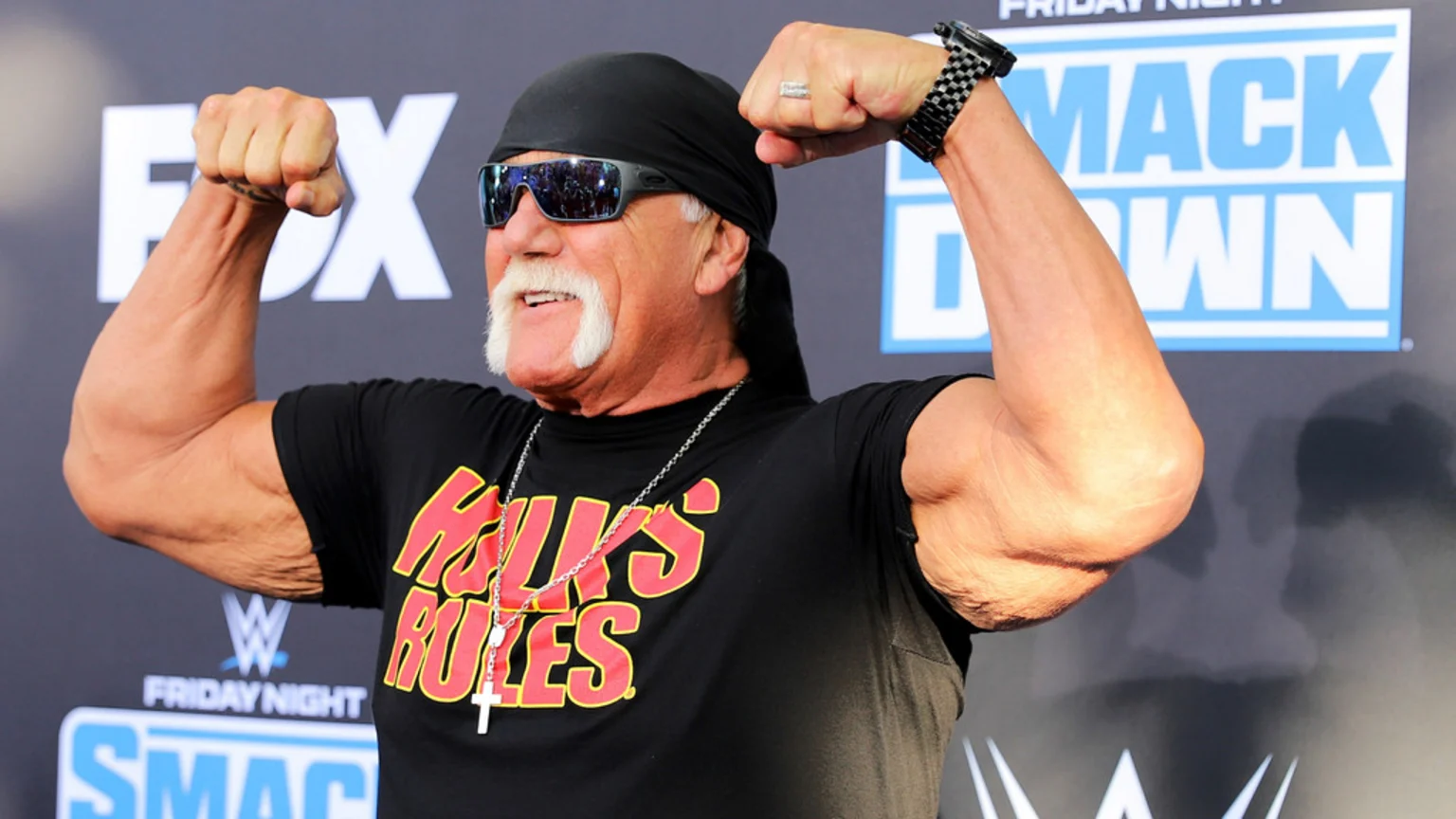 Hulk Hogan flexes his biceps while smiling, wearing a black bandana, sunglasses, and a "HULK'S RULES" t-shirt, with "FOX" and "SMACK DOWN" logos in the background.