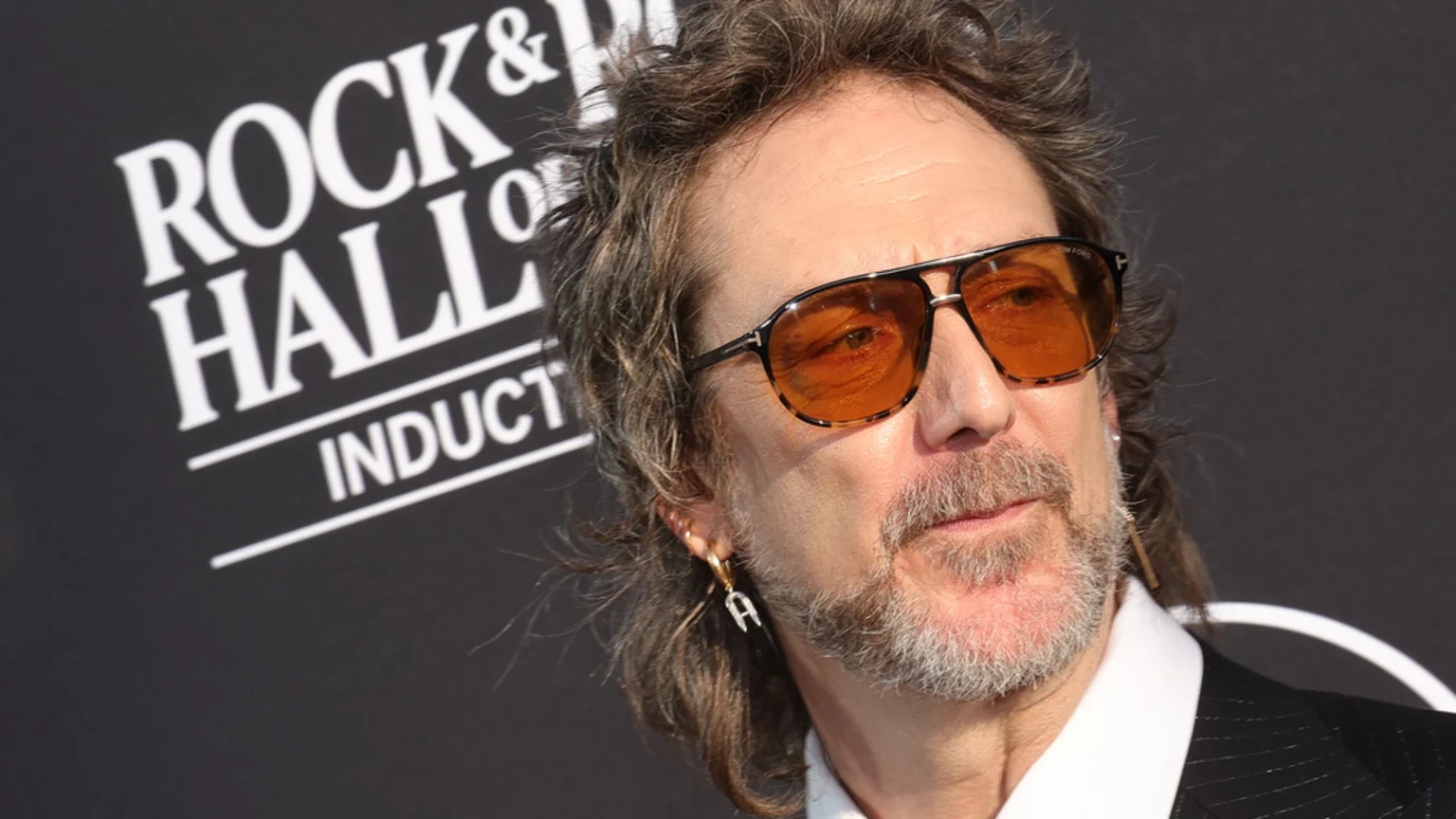 Close-up of a man with wavy hair, a beard, orange-tinted sunglasses, and an "A" earring, in front of a "ROCK & ROLL HALL OF INDUCTION" banner.