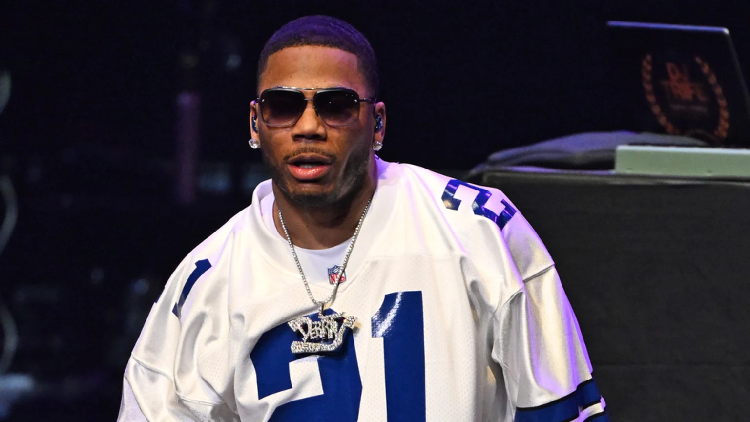 Nelly is shown from the chest up, wearing sunglasses, diamond stud earrings, a silver chain with diamond "DERRTY" and "91" pendants, and a white and blue football jersey with the number "91".
