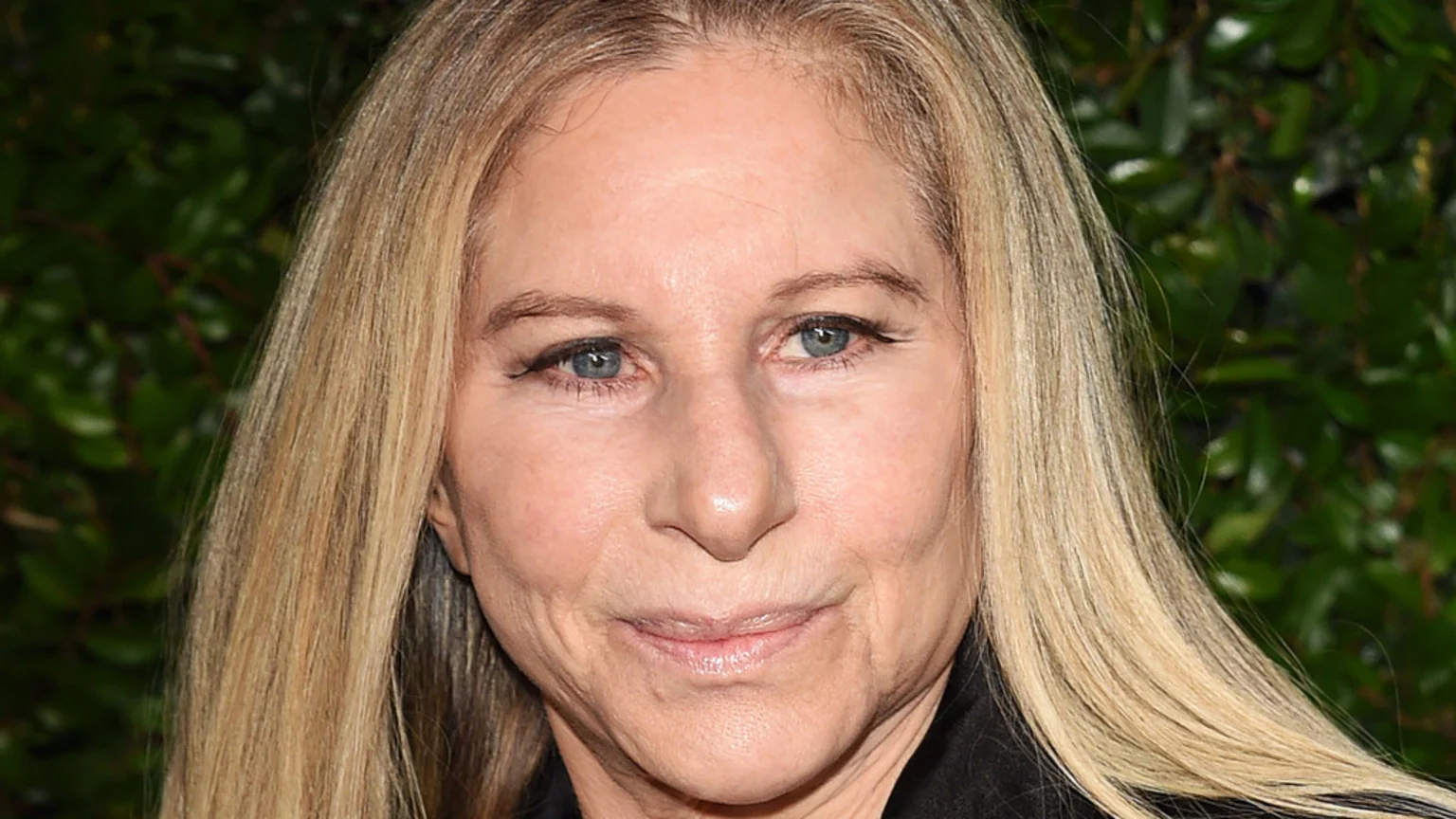 A close-up portrait of an older woman with long blonde hair and blue eyes, wearing black eyeliner, against a blurry green background.