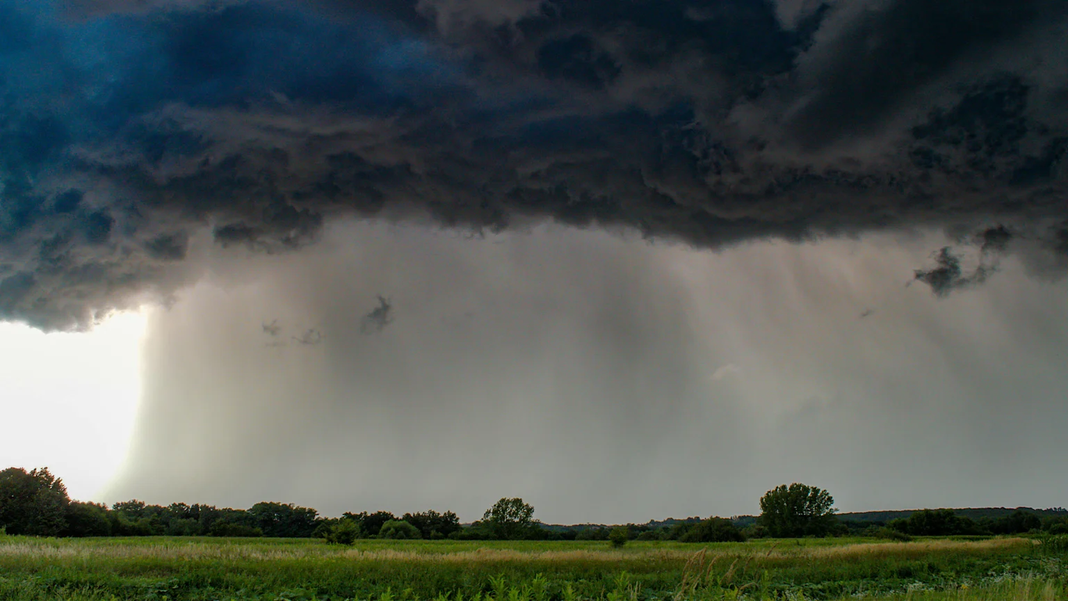 A dark, ominous thunderstorm sky with heavy clouds and visible rain falling over a green field and distant trees, with a bright flash of light on the left.