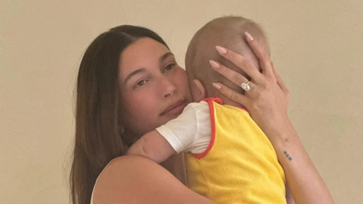 A woman with long brown hair, wearing a large oval diamond ring and a small tattoo on her forearm, holds a baby in a yellow top against a light beige background.