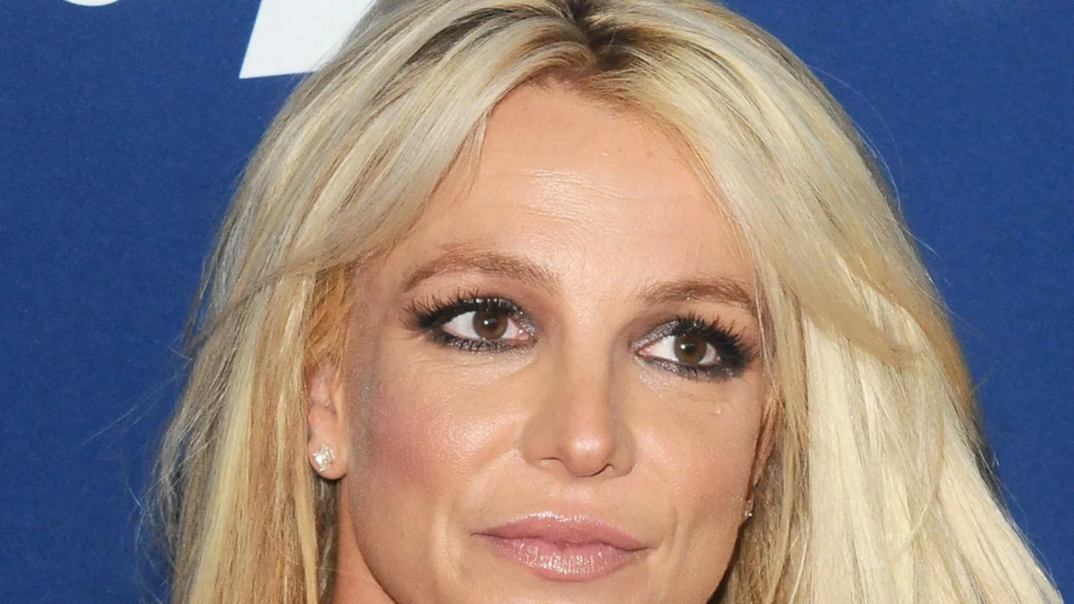 A close-up headshot of Britney Spears features her blonde hair, brown eyes with dark makeup, glossy light pink lips, and a stud earring, set against a solid blue background.