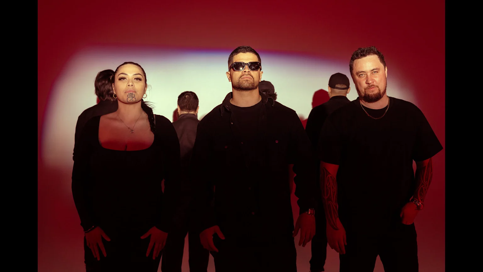 Three individuals—a woman with facial tattoos, a man in sunglasses, and another man with arm tattoos—stand wearing black in front of a red-lit background, with blurred figures behind them.