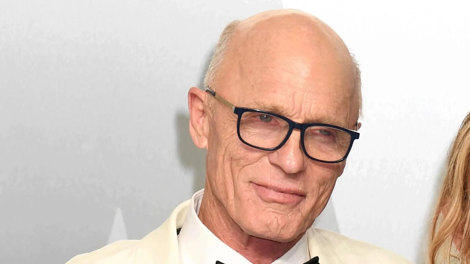 Ed Harris, a bald man in dark glasses, a light suit, and a bow tie, smiles slightly at the viewer.