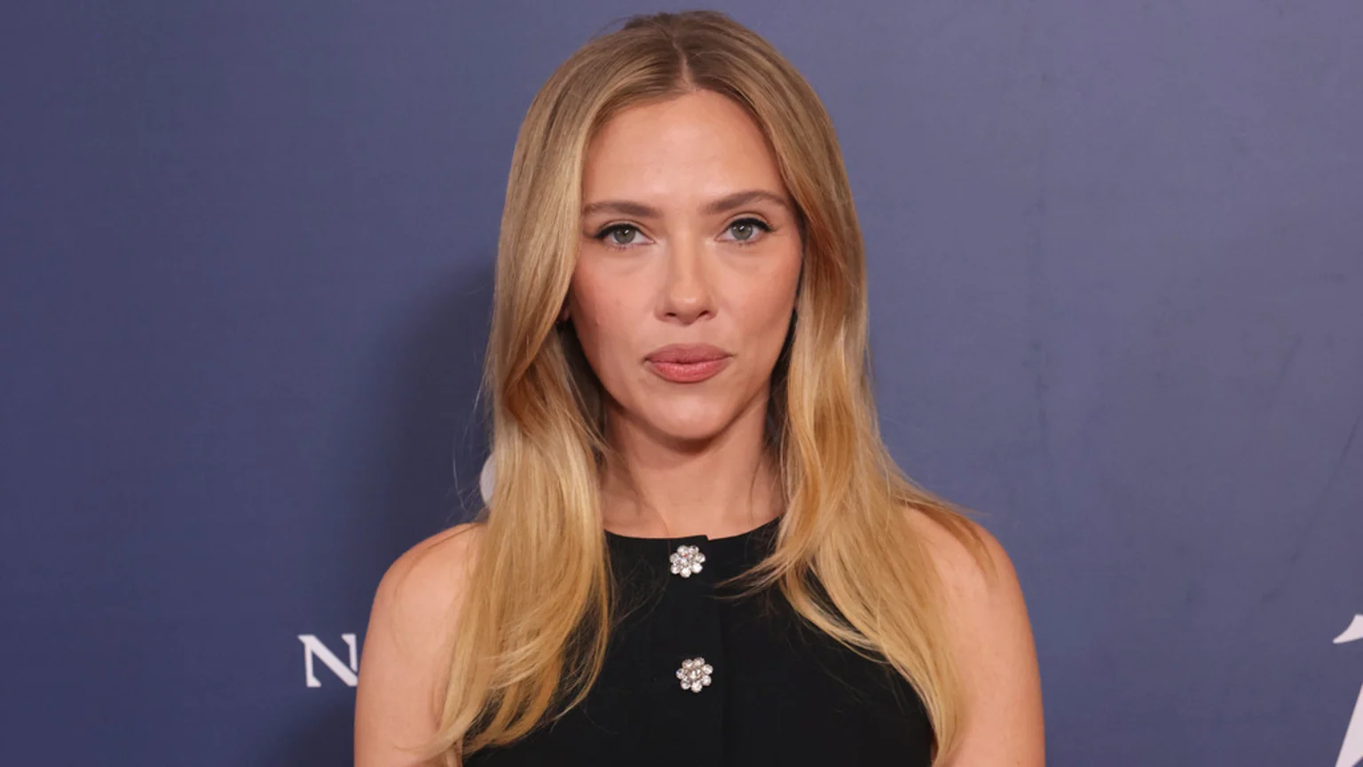 Scarlett Johansson, with long blonde hair and light makeup, wears a black top with two sparkly, floral buttons, looking directly at the camera against a blue background.