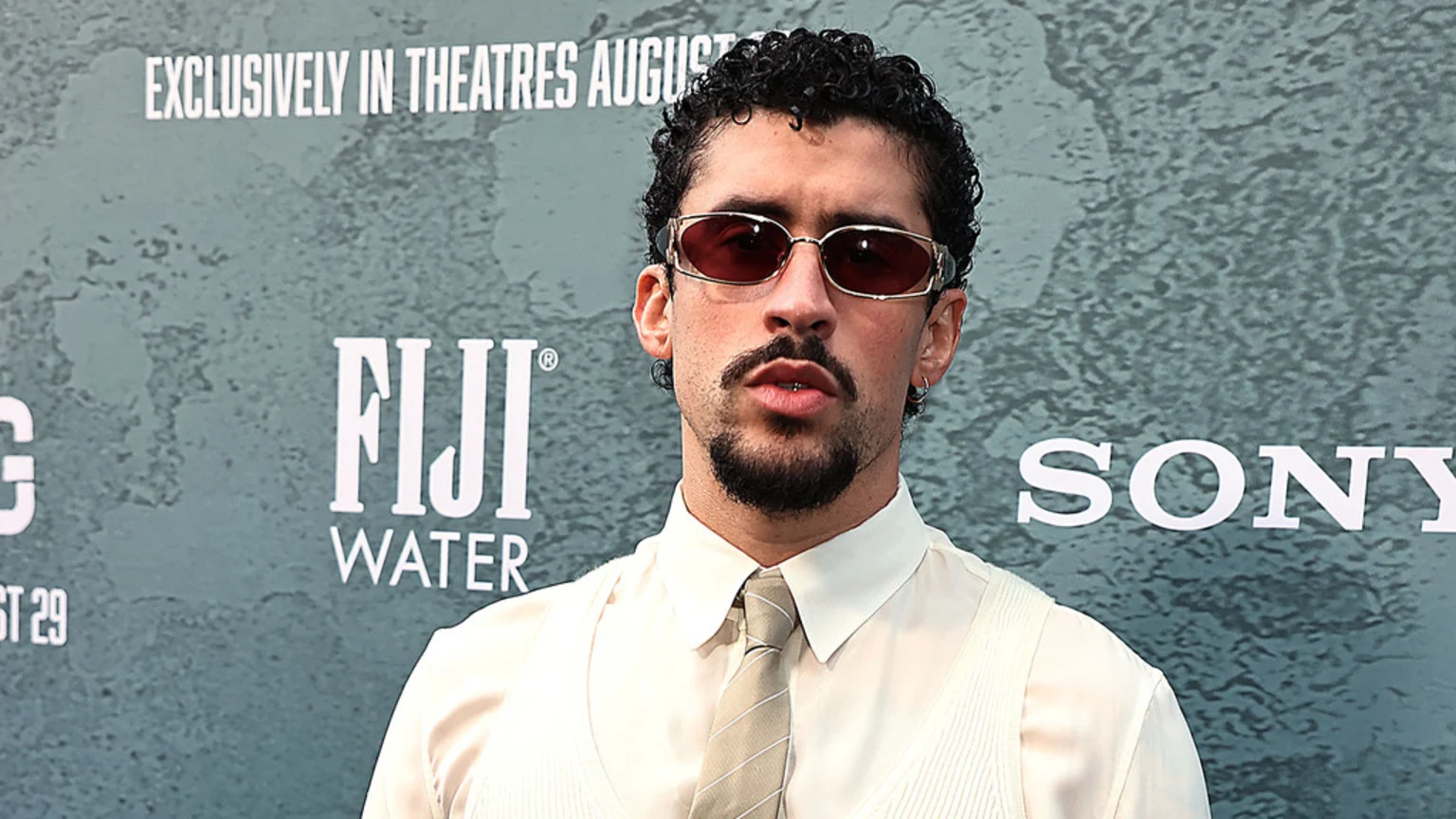 A man with curly hair, sunglasses, mustache, and goatee wears a shirt, tie, and vest in front of a textured wall with "FIJI WATER" and "SONY" logos.