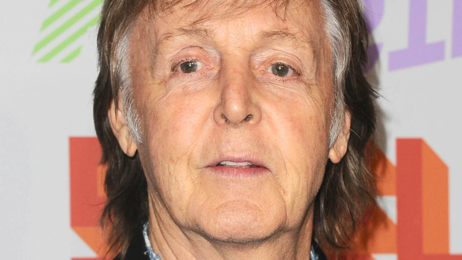 A close-up headshot of Paul McCartney, an older man with a neutral expression, prominent facial wrinkles, and long, grey-streaked brown hair, against a blurred background of colorful shapes.