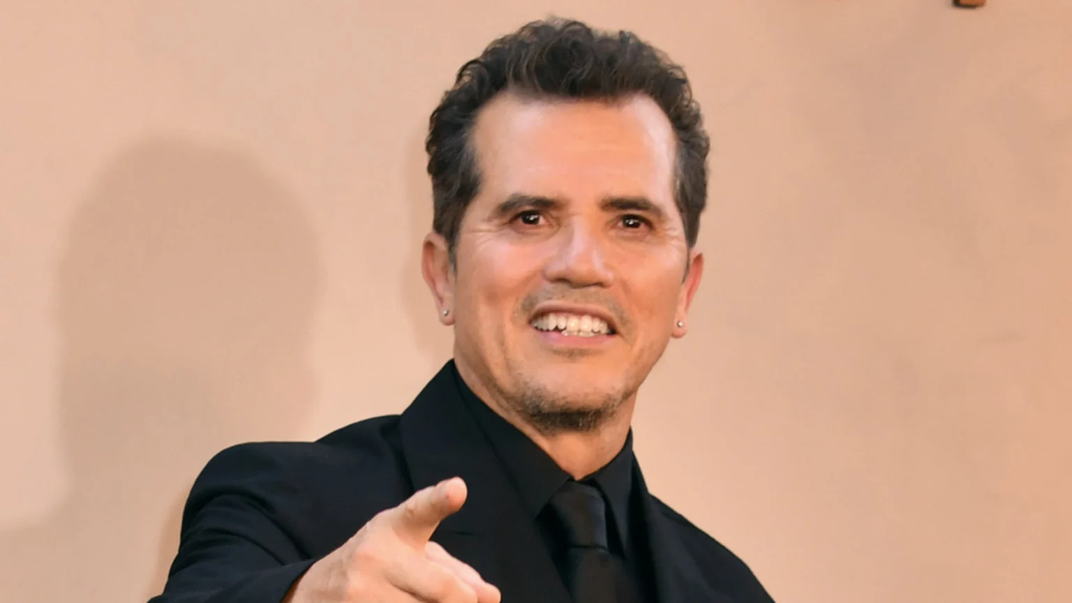 John Leguizamo, a man with dark wavy hair, black suit, and tie, wearing silver stud earrings, smiles and points forward against a plain light brown background.