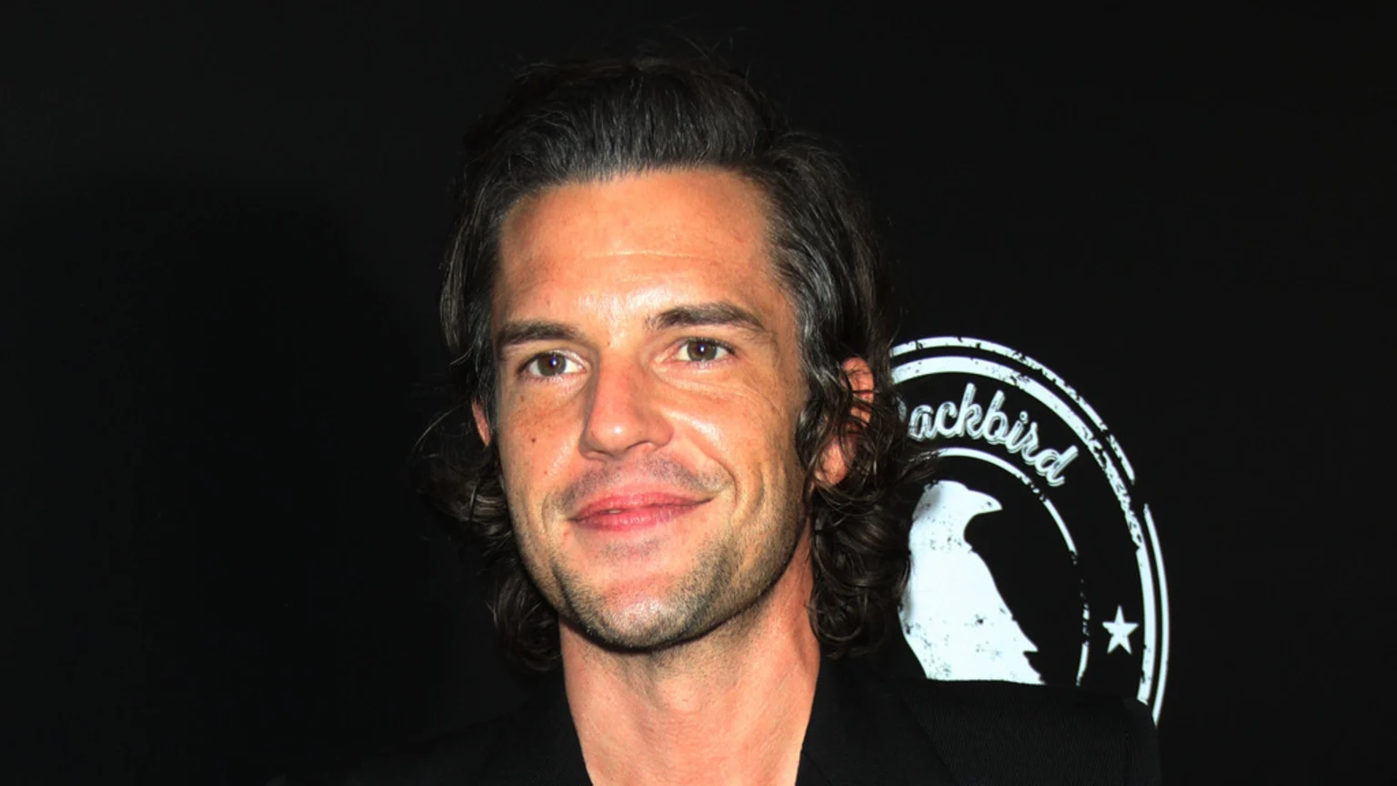 A portrait of musician Brandon Flowers with wavy dark hair, a light beard, and brown eyes, smiling slightly against a dark background with a partial white "ackbird" logo.