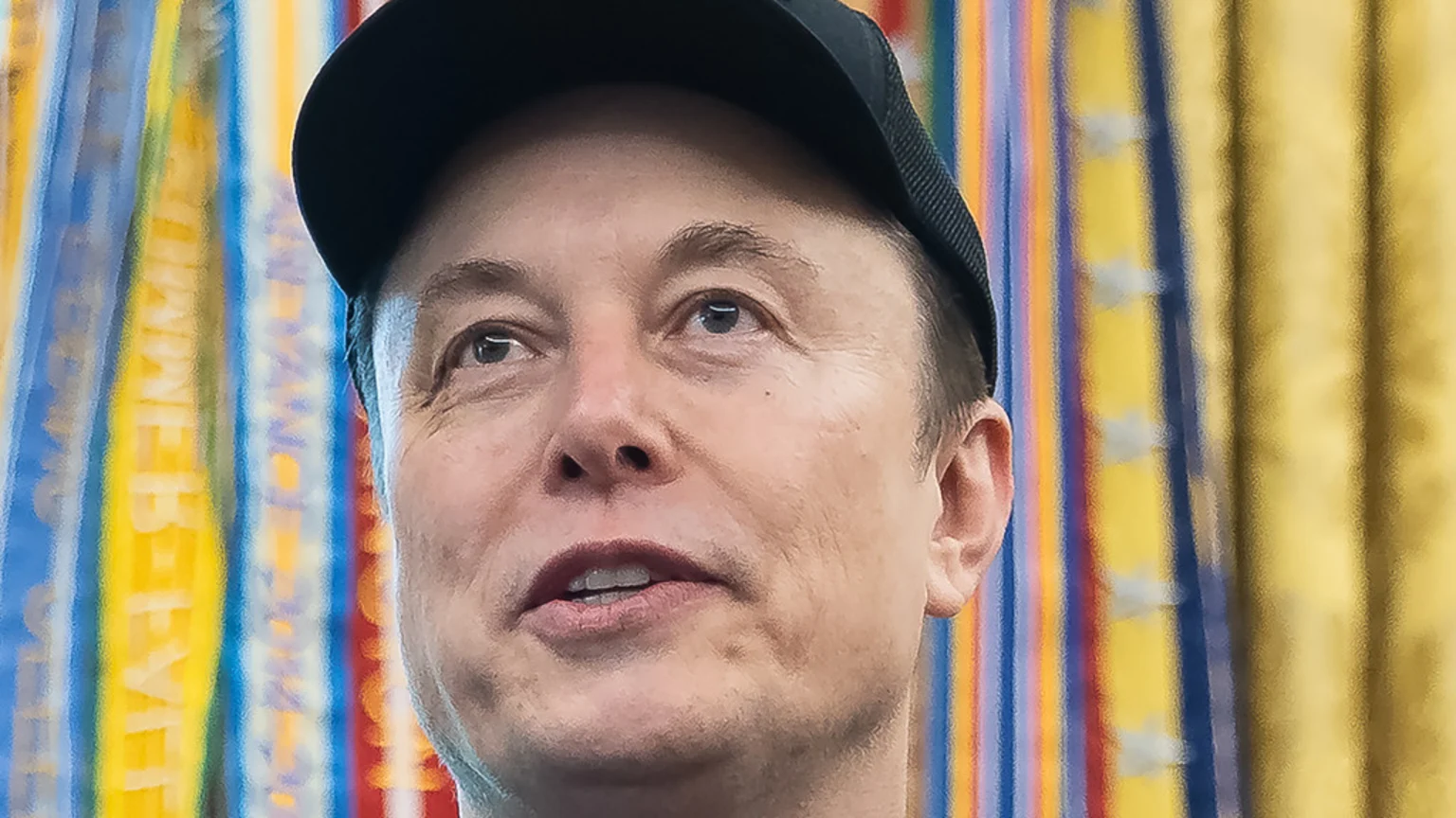 Close-up of Elon Musk wearing a black baseball cap, looking slightly up and to the right with his mouth slightly open, against a blurred, multicolored striped background.