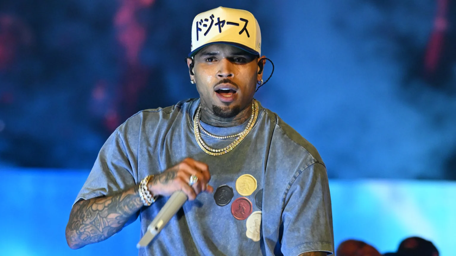 Chris Brown, sweating on stage, wears a light cap with Japanese text, a grey tee with circular patches, and multiple gold necklaces, holding a microphone.