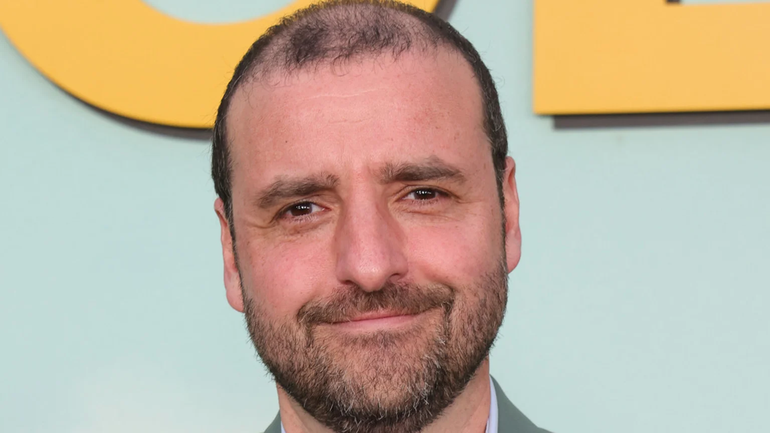 Close-up of a man with a receding hairline, dark beard, and a slight smile, wearing a light jacket against a light blue and yellow background.