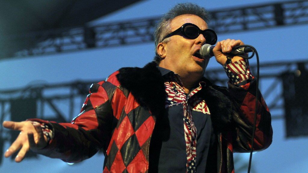 Jello Biafra, a man with grey hair and dark sunglasses, sings into a microphone, wearing a red and black checkered jacket with a fur collar and an American flag patterned shirt.
