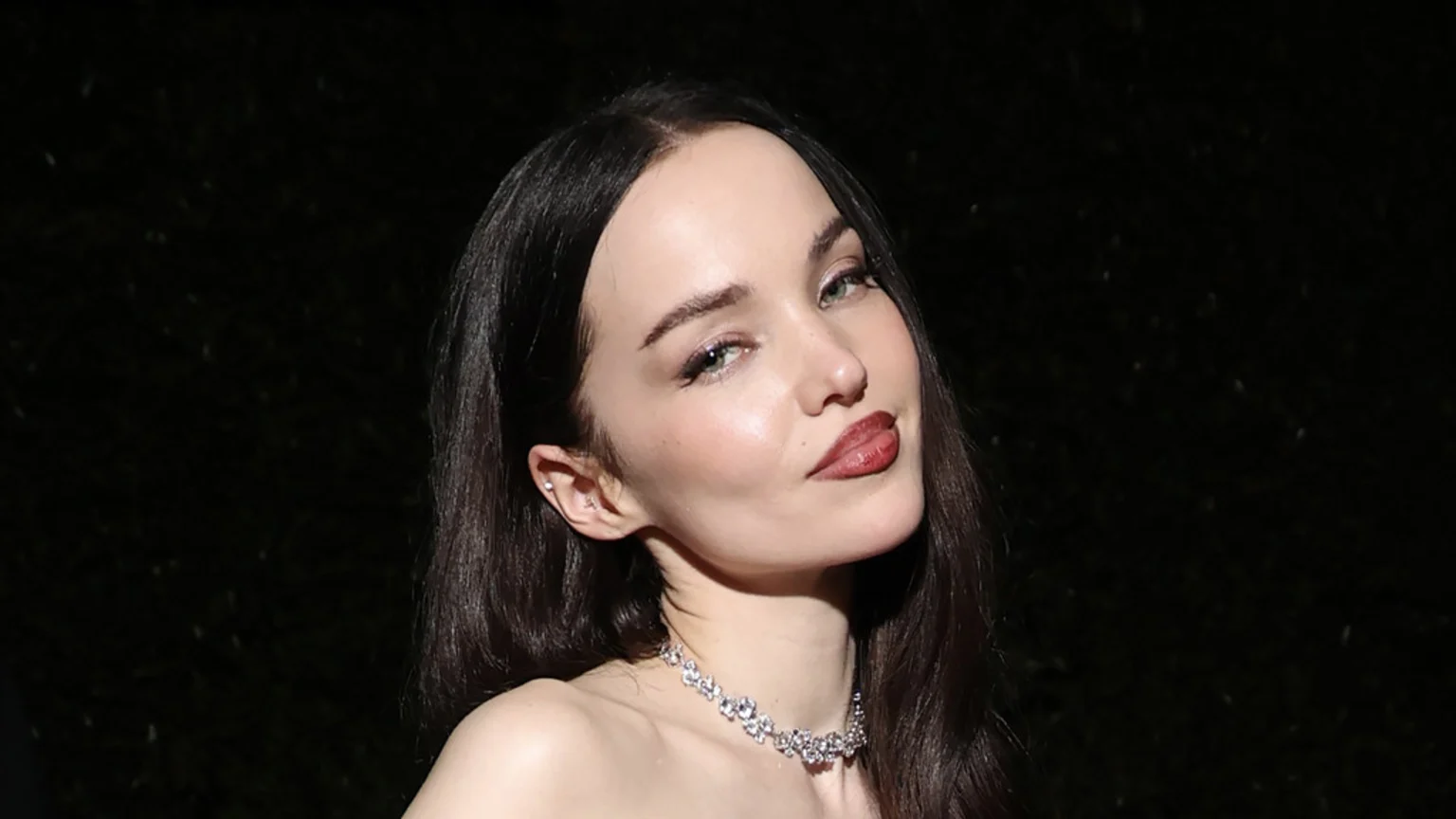 A close-up of a woman with long dark hair, light eyes, dark lipstick, and a sparkling choker necklace, looking at the viewer with a subtle smile.
