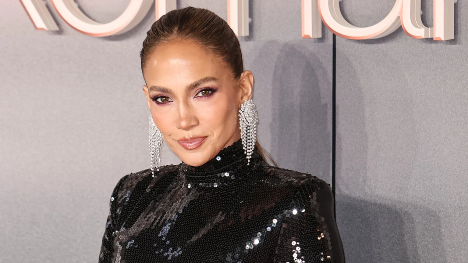 Jennifer Lopez, with her hair pulled back, smiles at the camera, wearing large sparkling chandelier earrings and a black sequined top with purple eyeshadow.