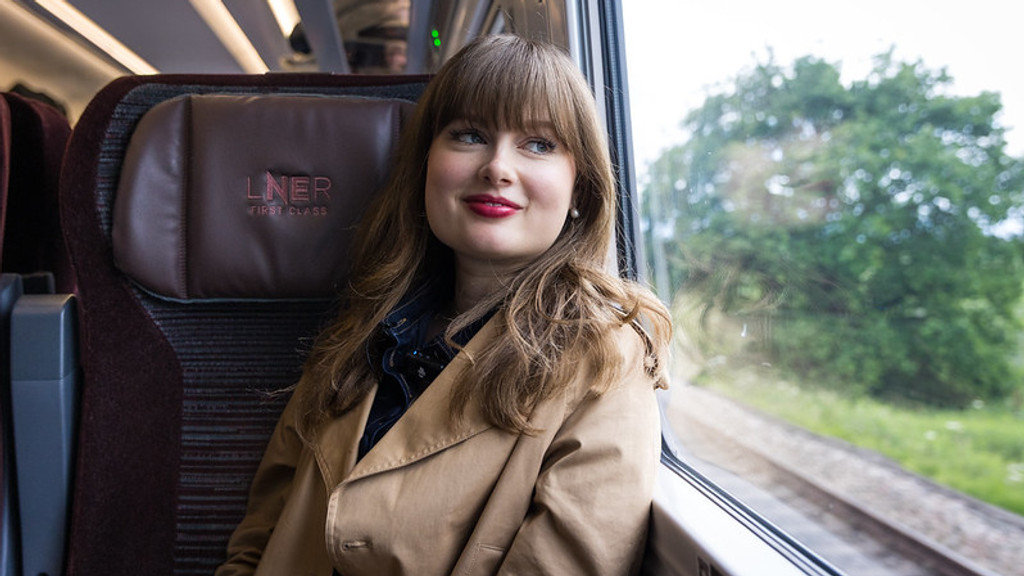 A young woman with long hair, red lipstick, and a tan trench coat smiles while looking out the window from an LNER First Class train seat as green scenery passes by.