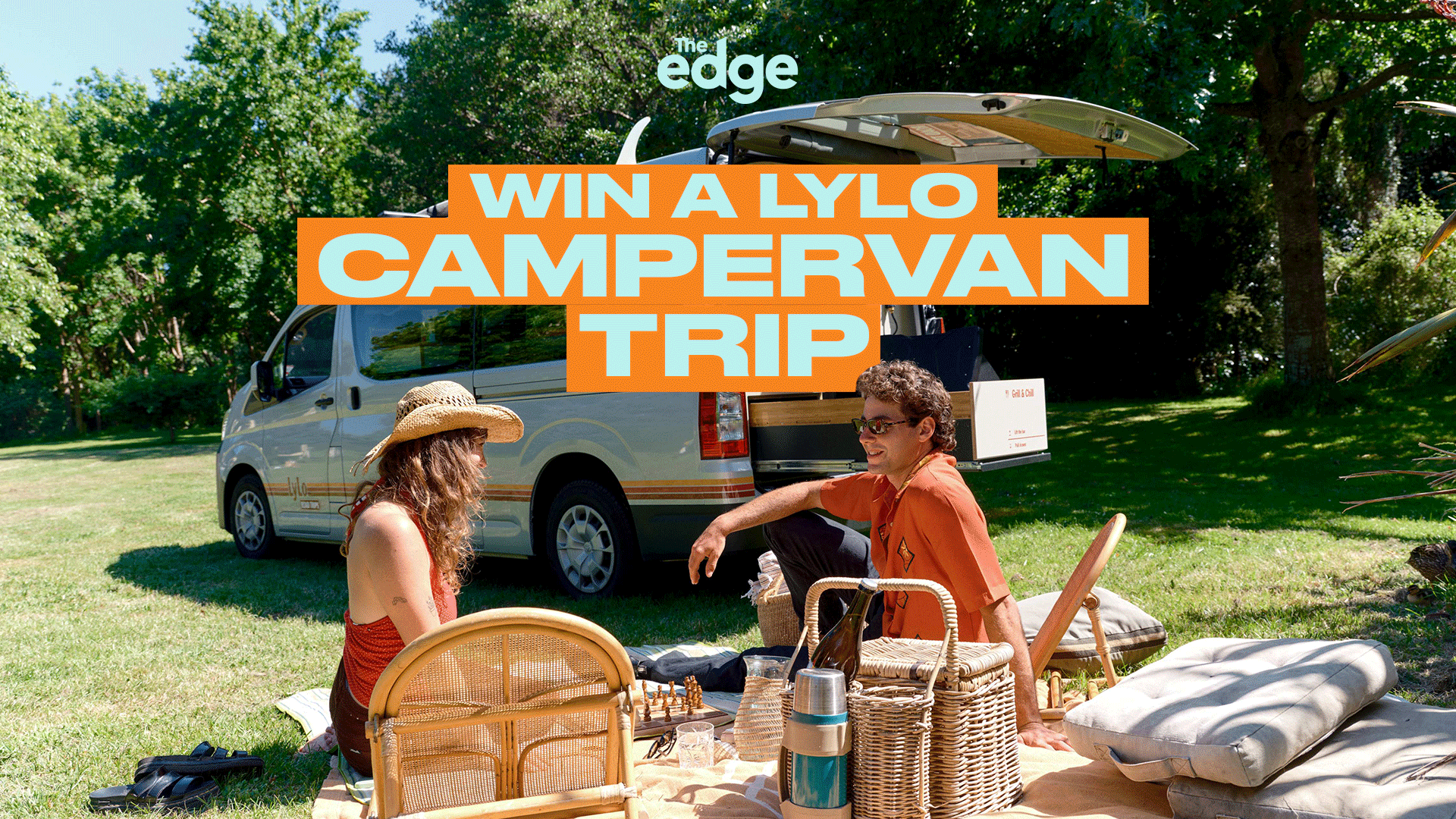A man and woman enjoy a sunny picnic on grass with a Lylo campervan parked behind them, surrounded by green trees.