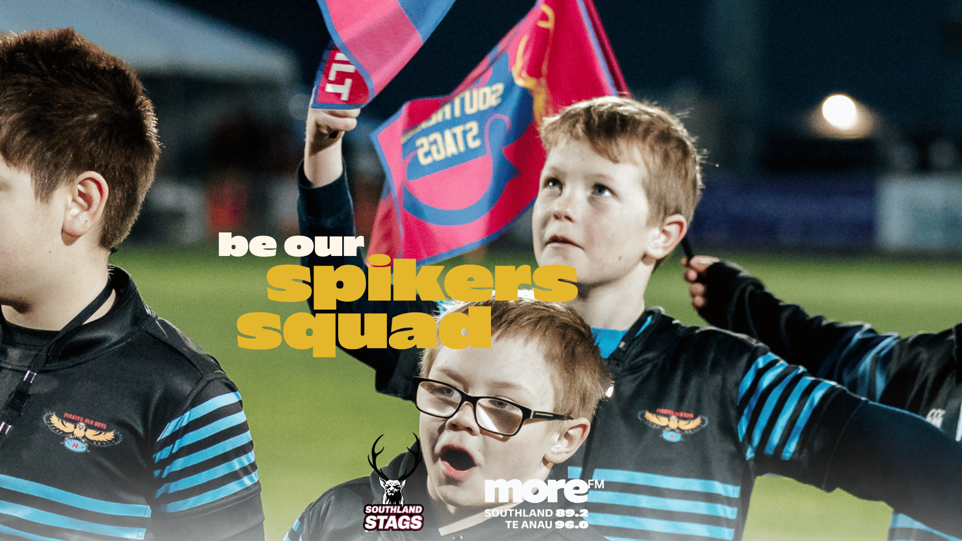Three young boys on a night sports field in team jerseys: one looks up holding two flags, another with glasses has his mouth open, and a third is visible from the side, with "be our spikers squad" text.