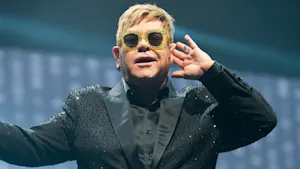 Elton John wearing yellow glasses and a black diamante suite - holding his hand to his ear left hand to his ear.