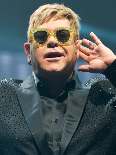 Elton John wearing yellow glasses and a black diamante suite - holding his hand to his ear left hand to his ear.