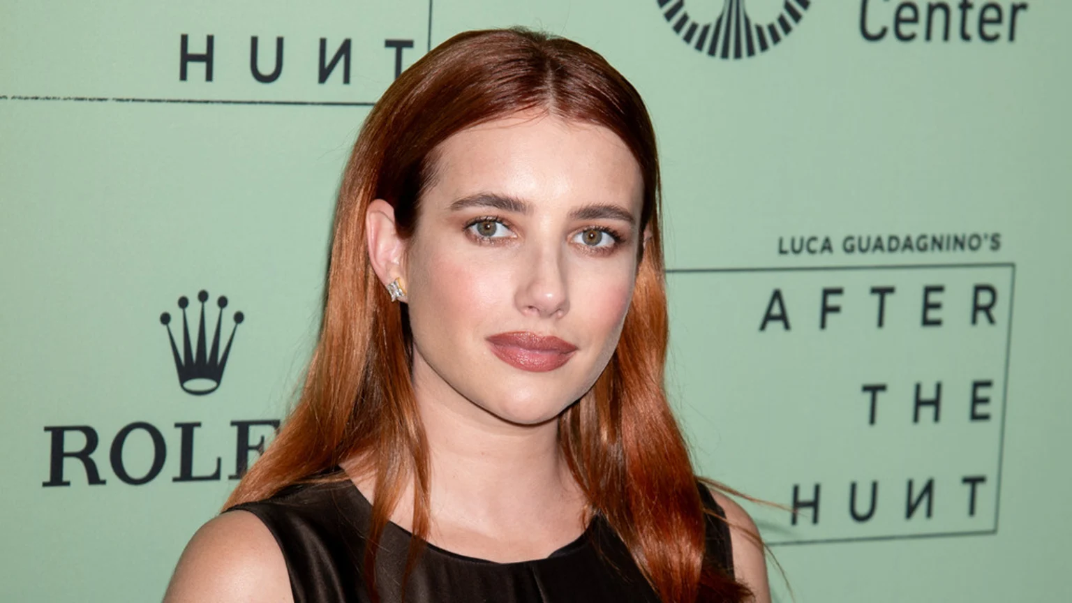 Emma Roberts, with long reddish-brown hair and a dark top, looks at the camera in a medium shot against a light green background with the Rolex logo and "After the Hunt" text.