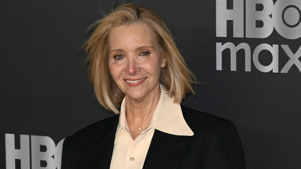 Lisa Kudrow, a blonde woman in a light collared shirt and dark blazer, smiles in front of a dark backdrop with the HBO Max logo.