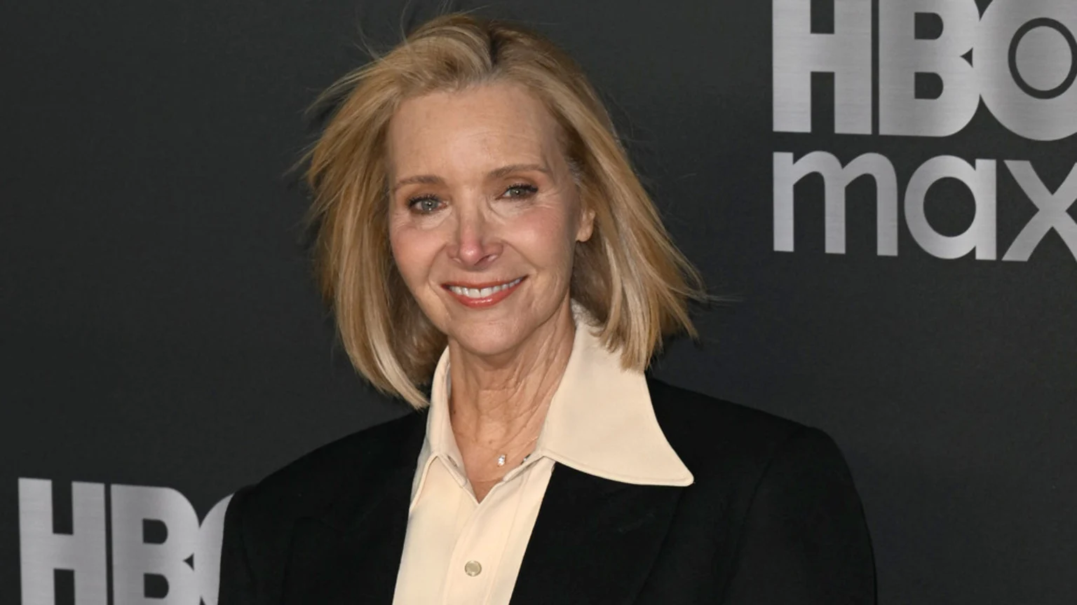 Lisa Kudrow, a blonde woman in a light collared shirt and dark blazer, smiles in front of a dark backdrop with the HBO Max logo.