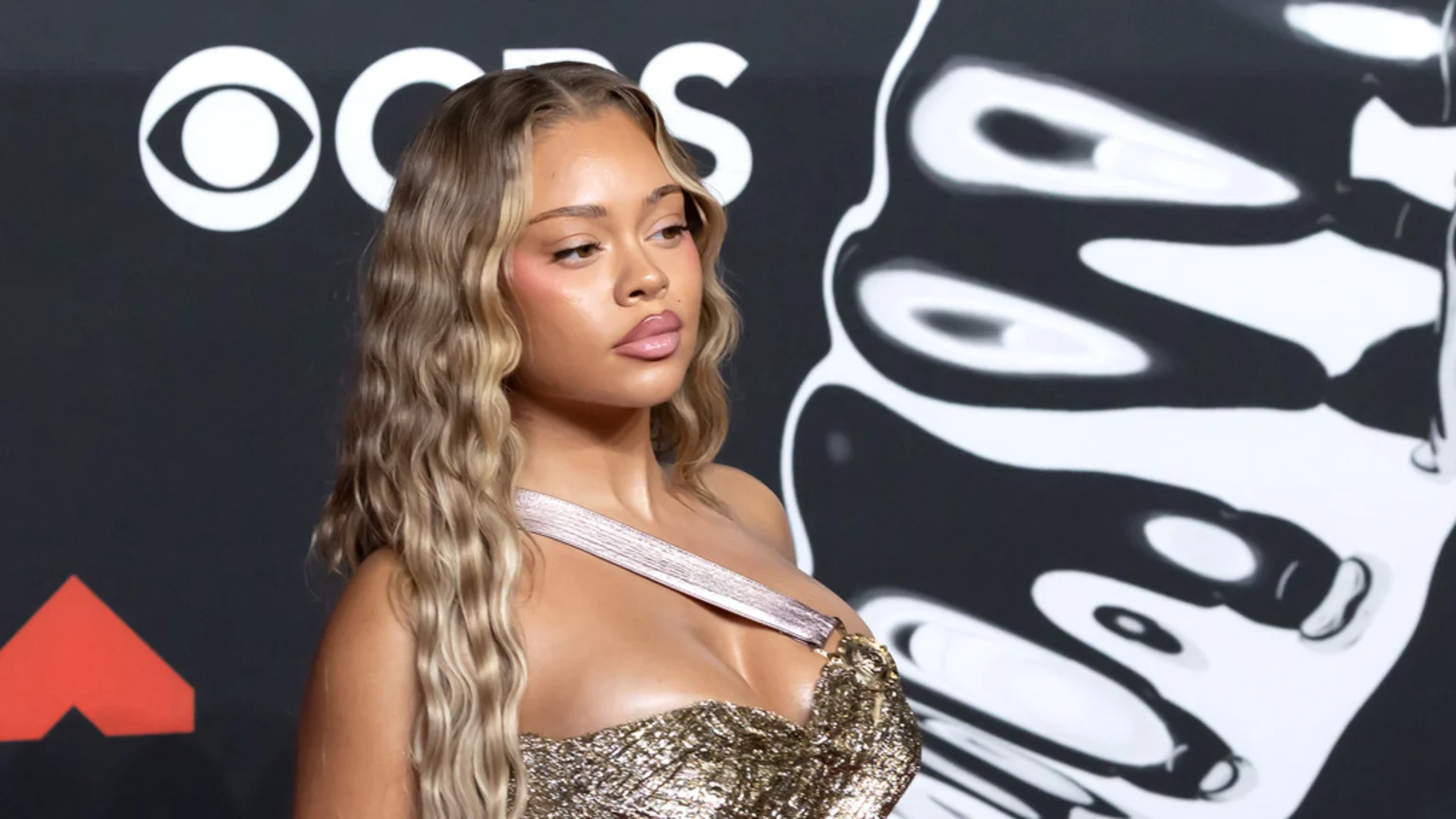 A woman with long, wavy light-brown hair and a shimmery gold top looks left, against a dark background with the CBS logo and abstract white patterns.
