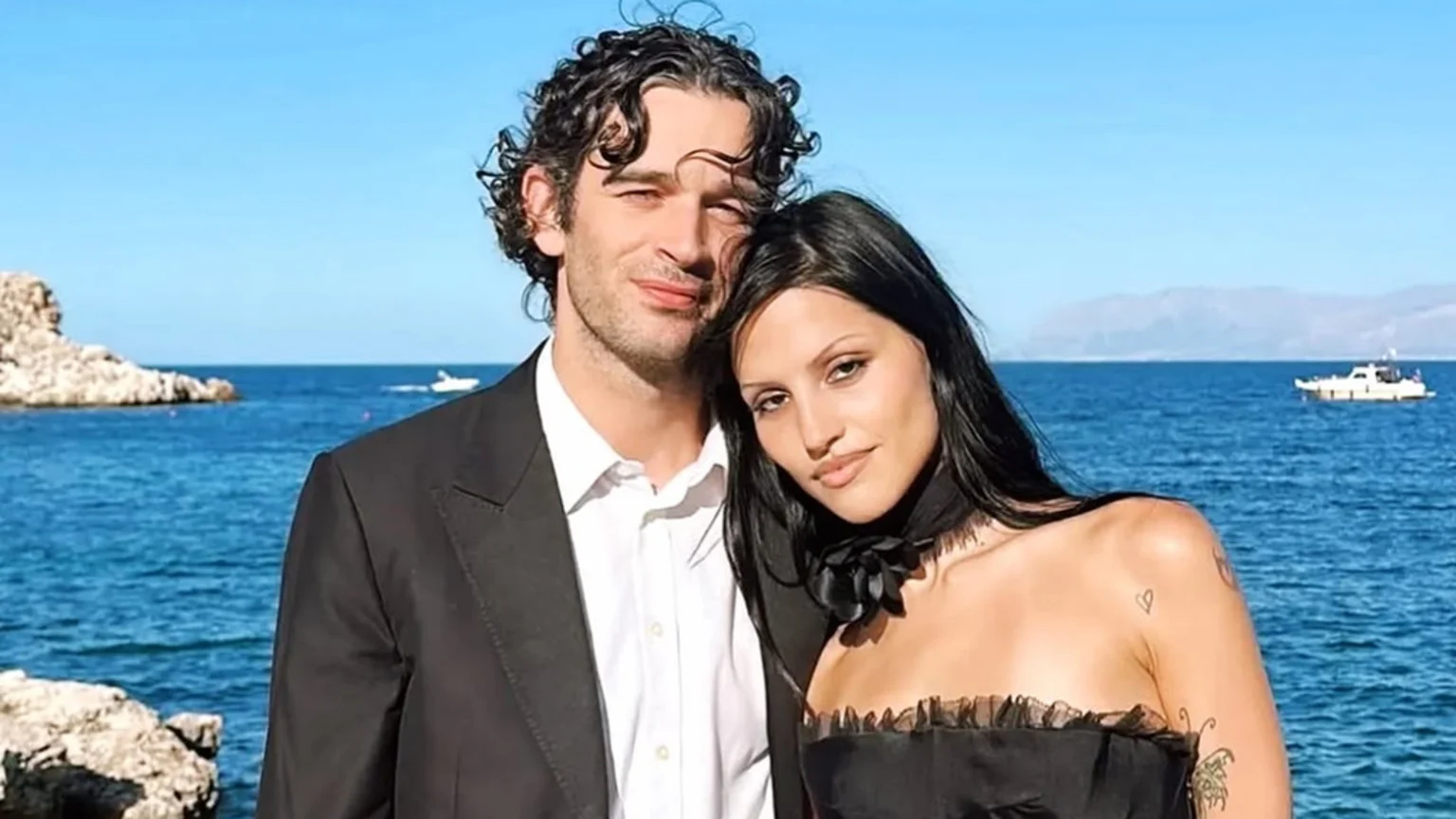 A man with curly hair in a suit and a woman with long dark hair in a black strapless top pose by the sea on a sunny day.