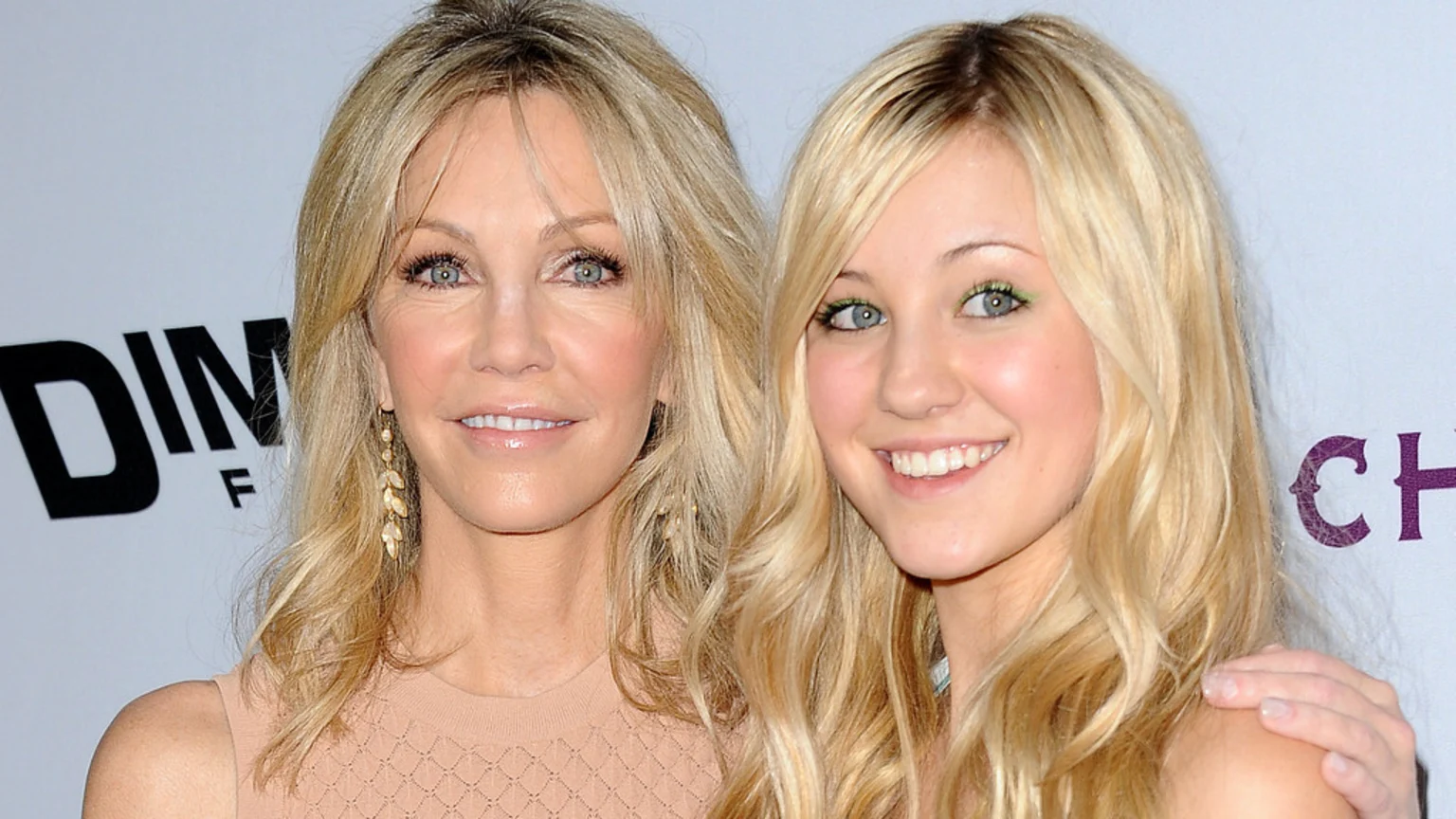 Two smiling blonde women, one older and one younger, both with blonde hair, pose together looking at the camera, with the older woman's hand on the younger woman's shoulder.