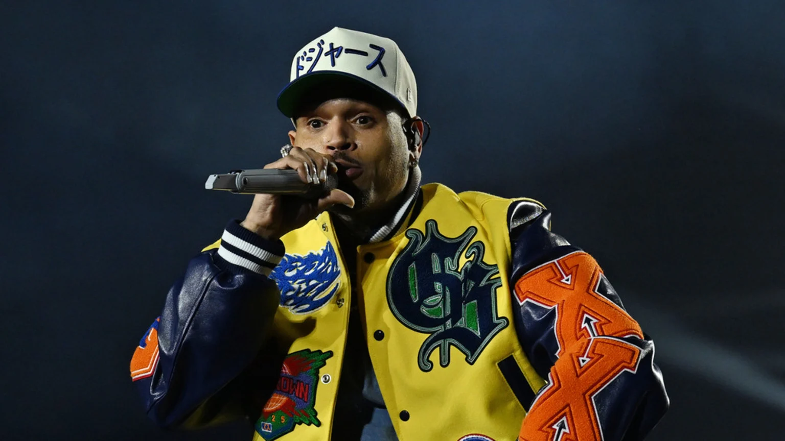 Chris Brown on stage, wearing a yellow varsity jacket with blue sleeves and a white cap with Japanese text, singing into a microphone.
