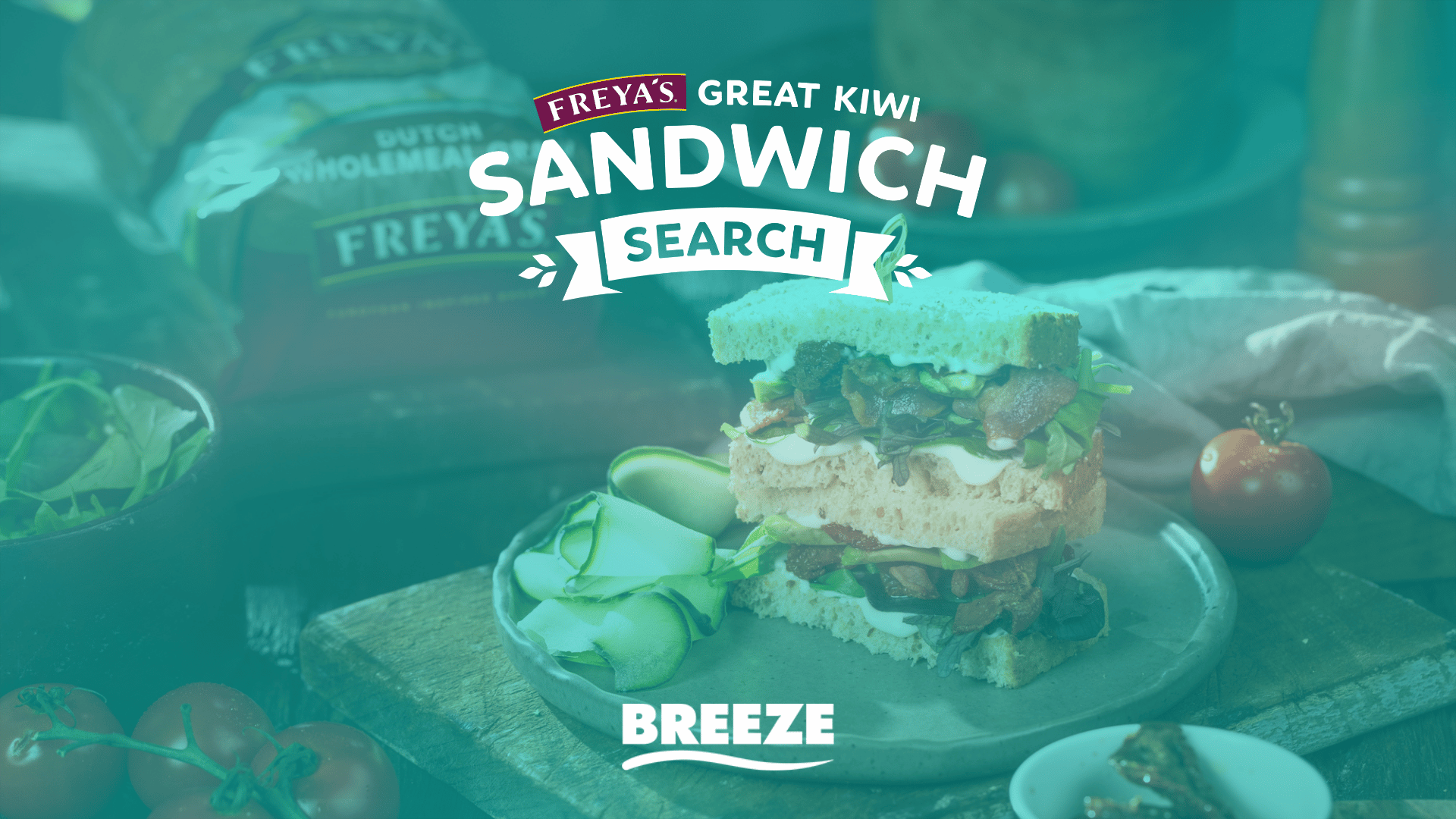 A layered sandwich, cucumber, and vegetables are on a wooden surface with a loaf of Freya's bread in the background, overlaid with "FREYA'S GREAT KIWI SANDWICH SEARCH" and "BREEZE" text.