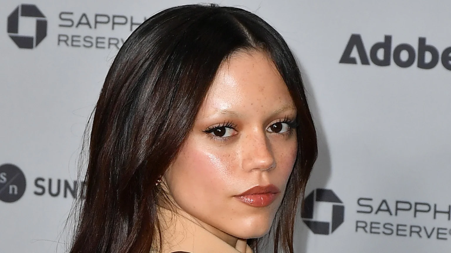 A close-up portrait of Jenna Ortega with dark hair, light eyebrows, freckles, and glossy lips, against a light background with blurred logos.