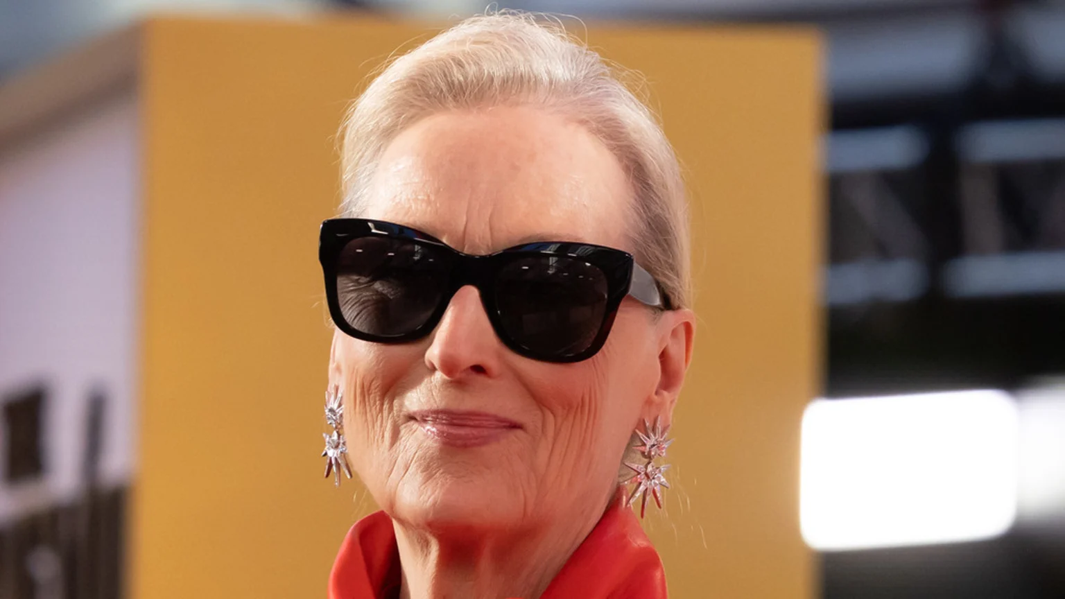 Meryl Streep smiles faintly, wearing large black sunglasses, a red top, and sparkly star-shaped earrings, with her light hair pulled back against a blurred golden background.