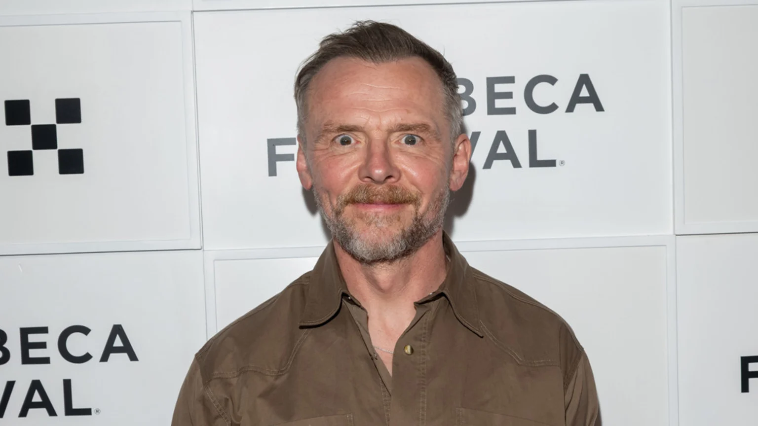 Actor Simon Pegg, with a beard and short hair, stands against a white wall with "TRIBECA FESTIVAL" text, looking at the camera with wide eyes and a slight smile.