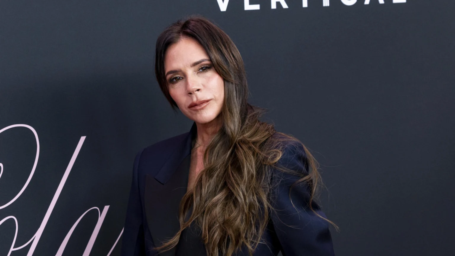 Victoria Beckham, with long wavy brown hair, poses in a dark blazer with light makeup, looking at the camera against a dark background with white text.