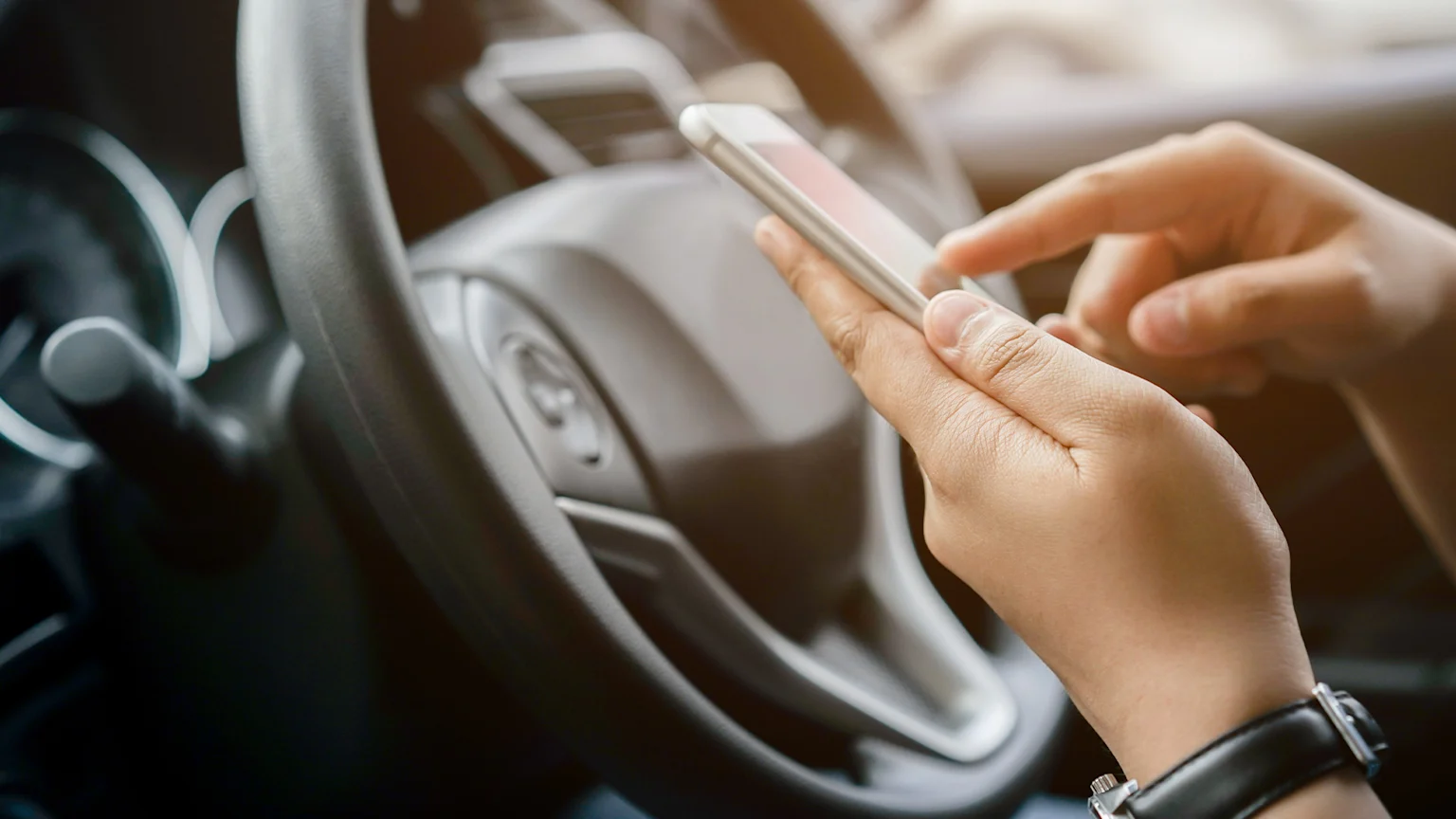 Woman's hand holding phone in front of steering wheel of a car.