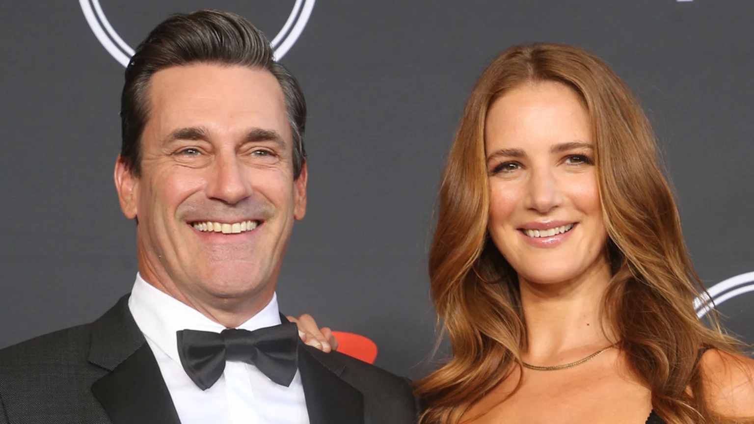 A smiling man in a suit and bow tie stands next to a smiling woman with long brown hair, who has her hand on his arm, against a dark background.