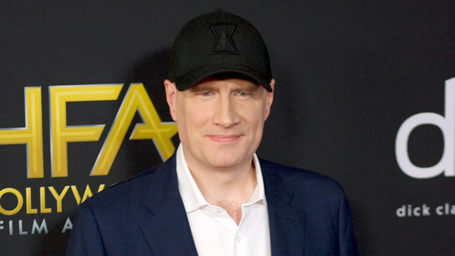 Kevin Feige smiling, wearing a black cap with a Black Widow logo, a dark suit, and a white shirt, against a dark background with "HFA" and "dick cla" visible.