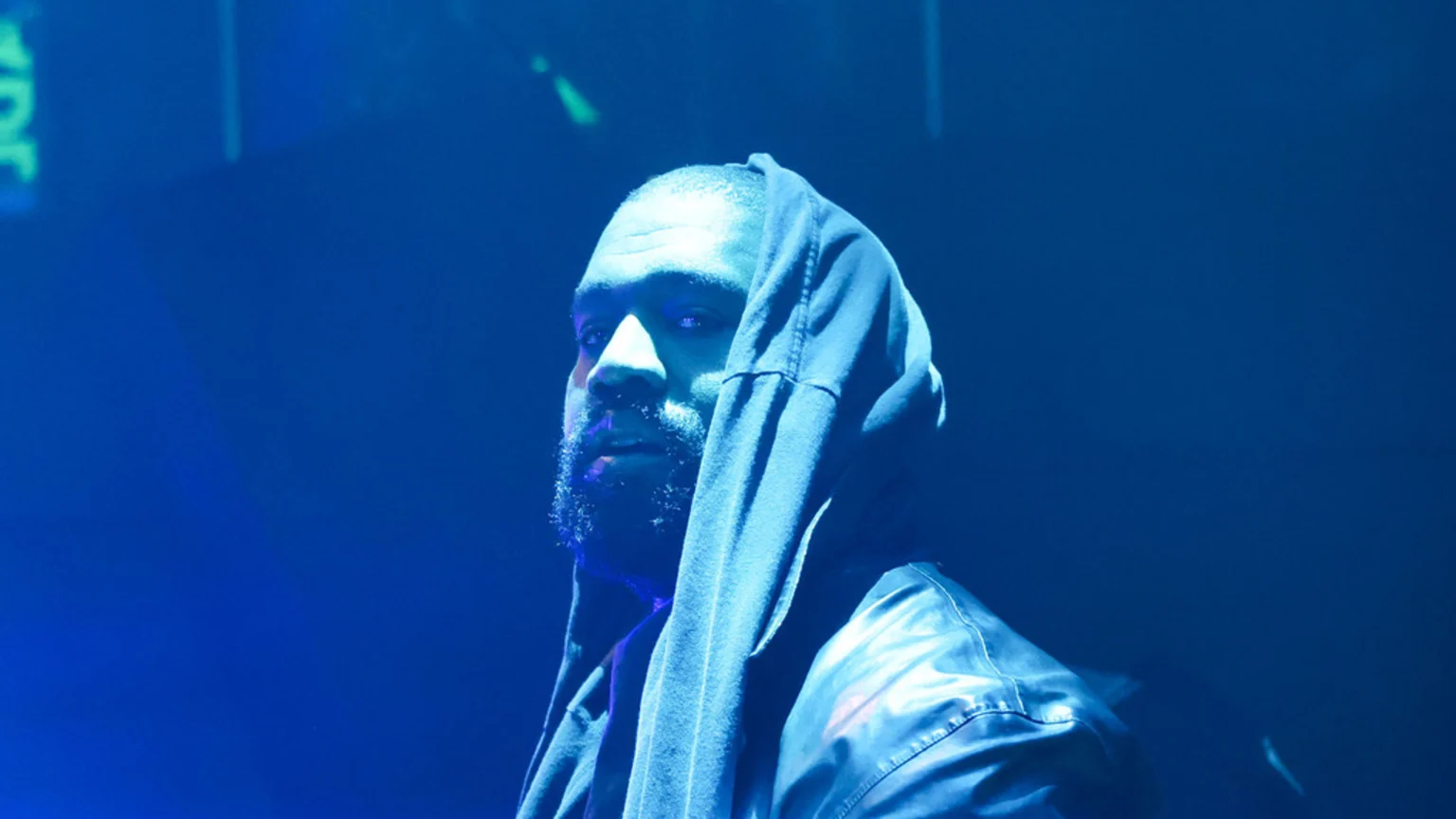 A bearded man with a serious expression, wearing a dark hooded top and jacket, looks left under intense blue light, with his eyes appearing to glow white.