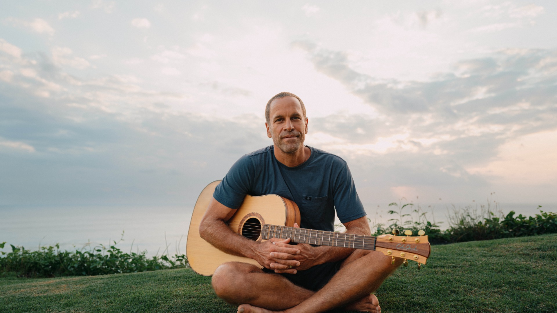 A man sits cross-legged on green grass, holding an acoustic guitar, with the ocean and a cloudy sky in the background.